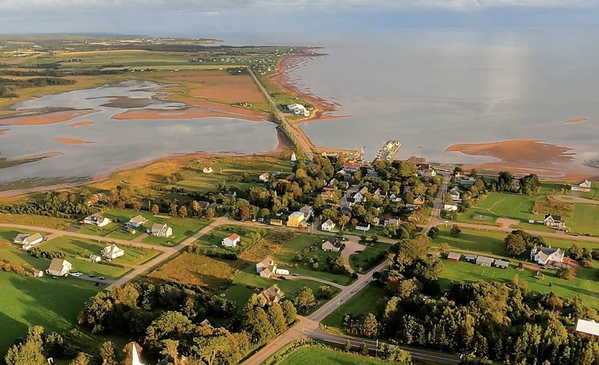Aerial view of Victoria-by-the-Sea showing the picturesque village with its historic buildings, long wooden wharf, red coastline, and calm protected waters ideal for paddle boarding